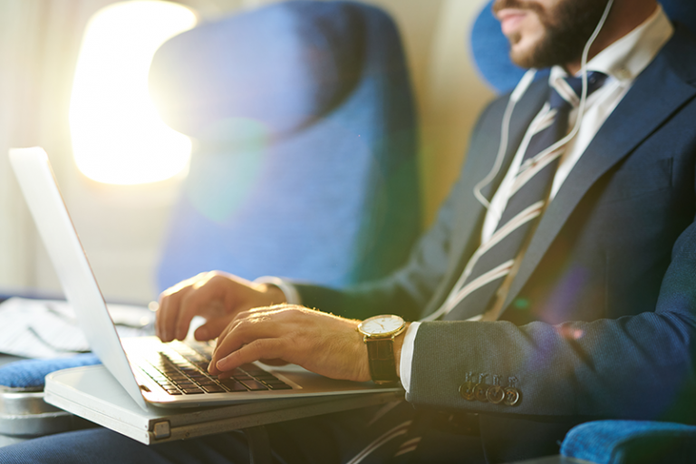 5 Crucial Cybersafety Tips for Business Travelers - Man wearing a suit and earbuds is visible from the waist up to his nose is on an airplane, working on a laptop