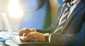 5 Crucial Cybersafety Tips for Business Travelers - Man wearing a suit and earbuds is visible from the waist up to his nose is on an airplane, working on a laptop