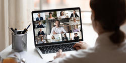 Over-the-shoulder image of a person sitting at a desk with a cup of tea to watch a meeting on their laptop, which shows a grid of several video feeds on screen